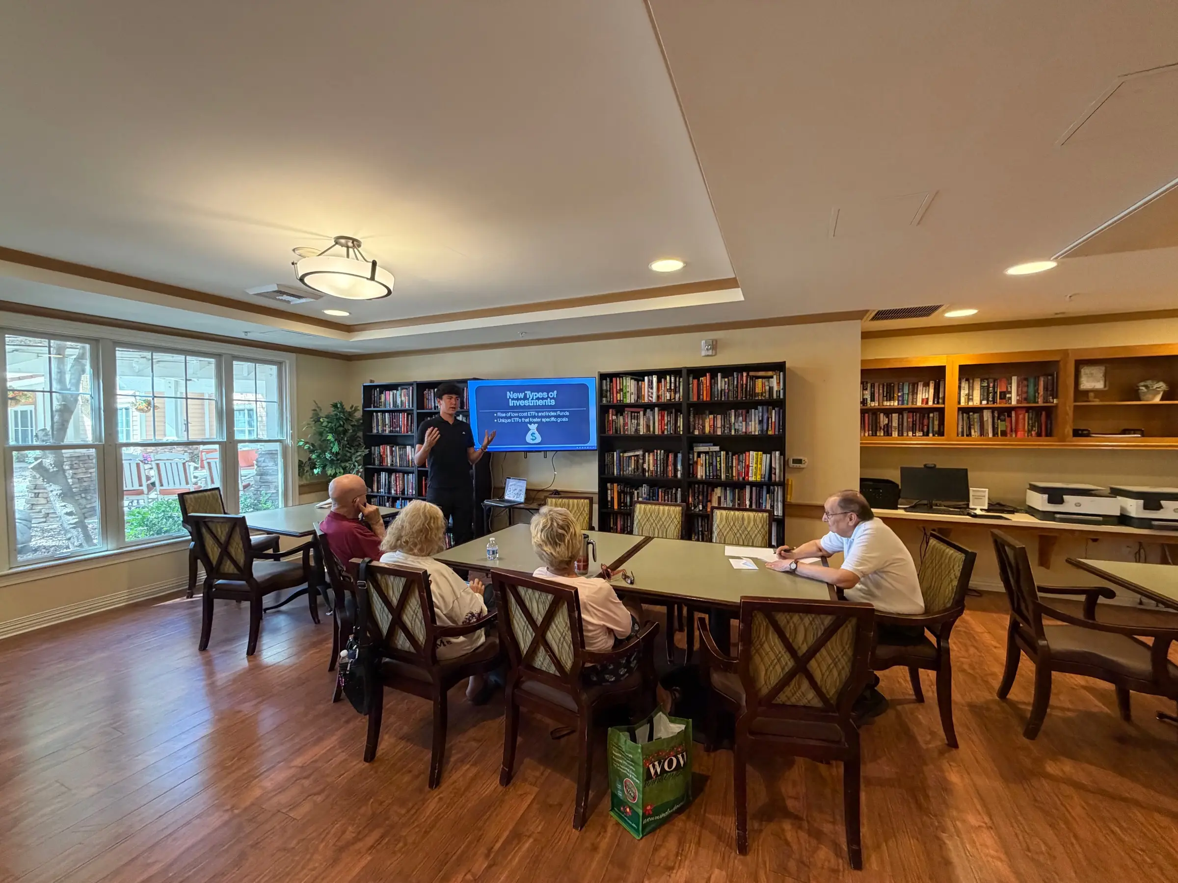 A session under way in a community library: a student volunteer standing beside a screen, with a small group of older adults seated around tables next to a large window.