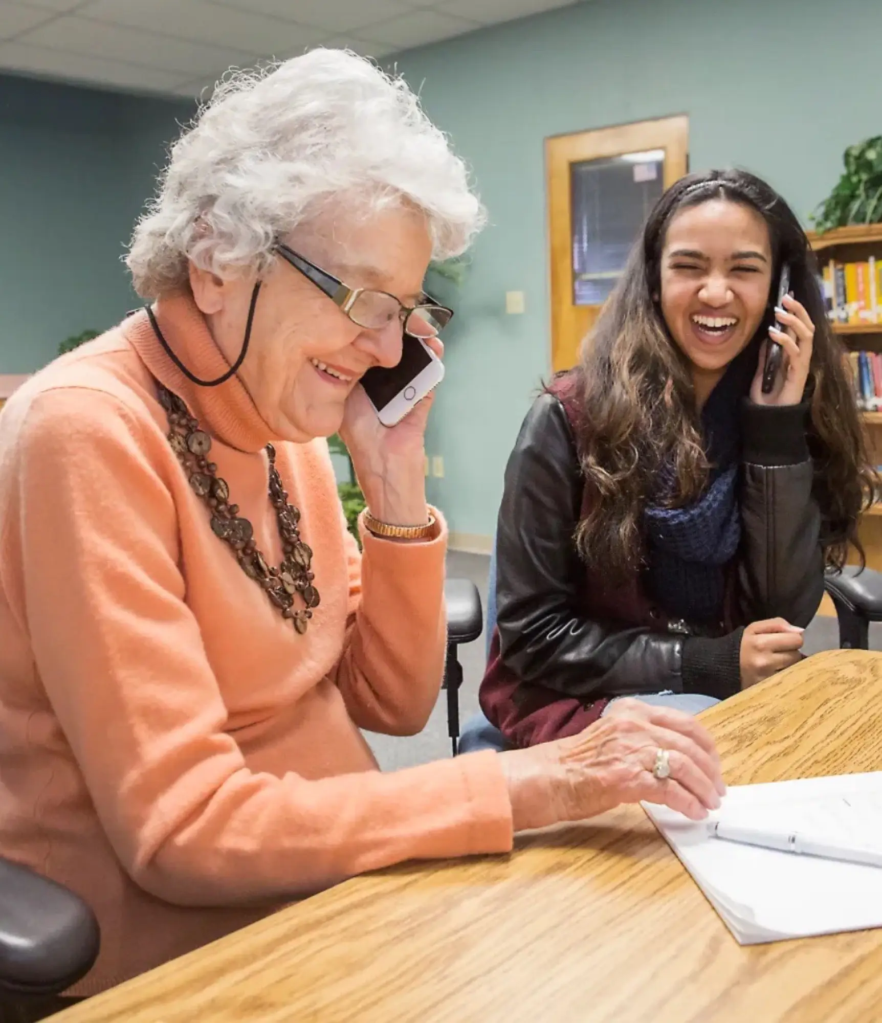 An older woman and a young woman sitting together at a table, both on their phones and laughing.